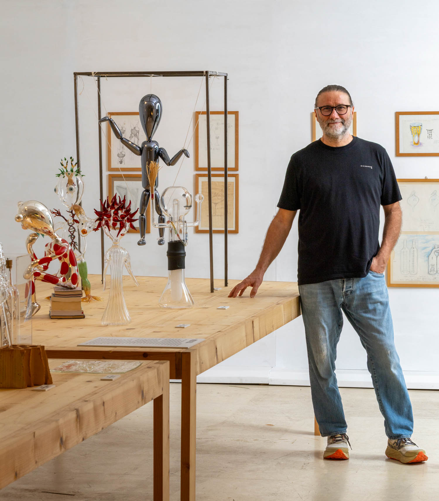 A man in a black t-shirt and jeans stands next to a wooden table displaying various glass sculptures and a metallic humanoid figure.