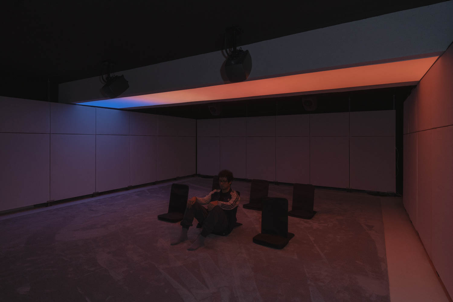 A person sits on the floor in a dim, minimalist room illuminated by blue and orange ambient light, surrounded by several floor cushions.