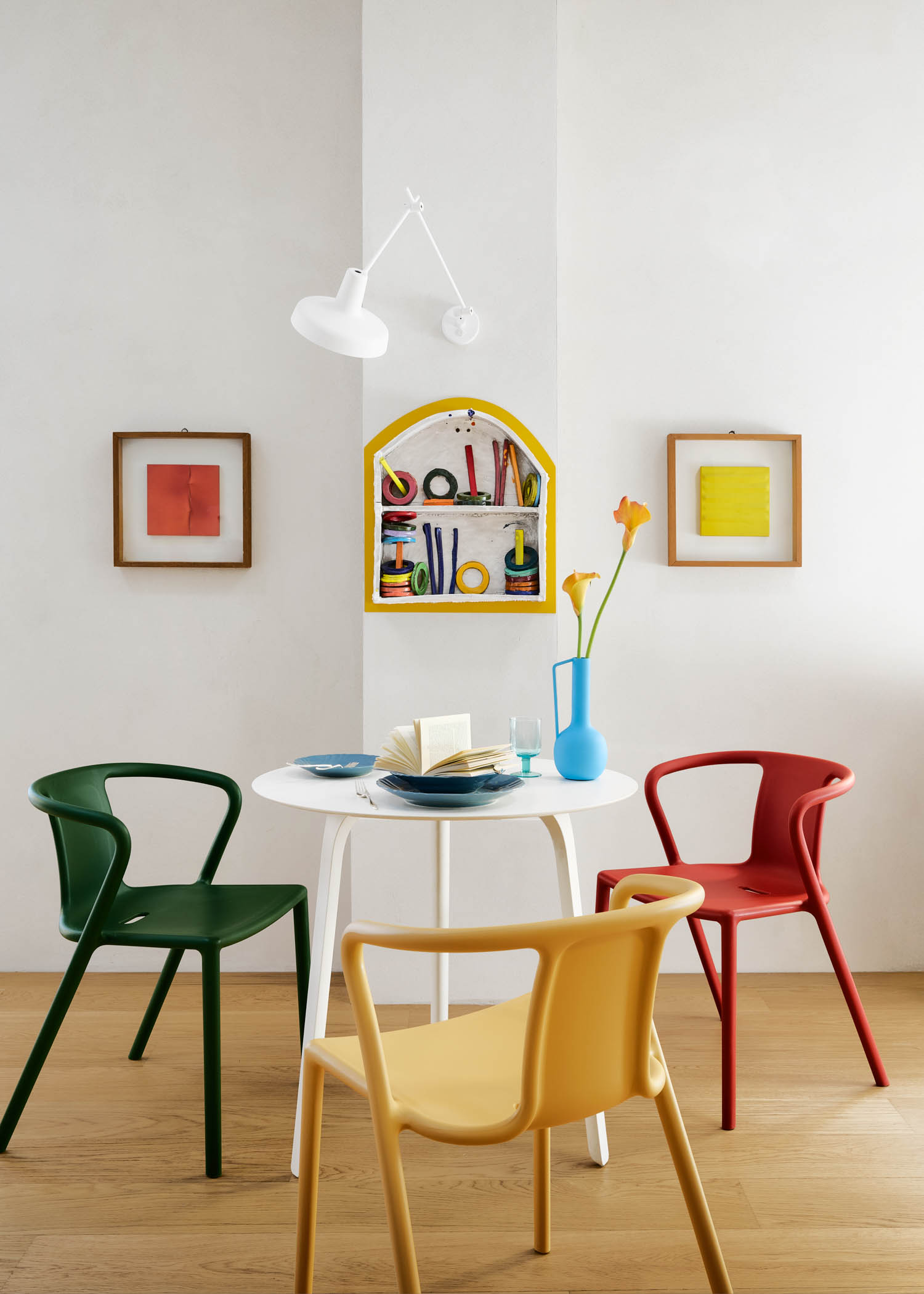A white dining table with an open book and a blue vase of flowers, surrounded by three colorful plastic chairs in front of a wall with two framed art prints and a decorative yellow wall shelf.