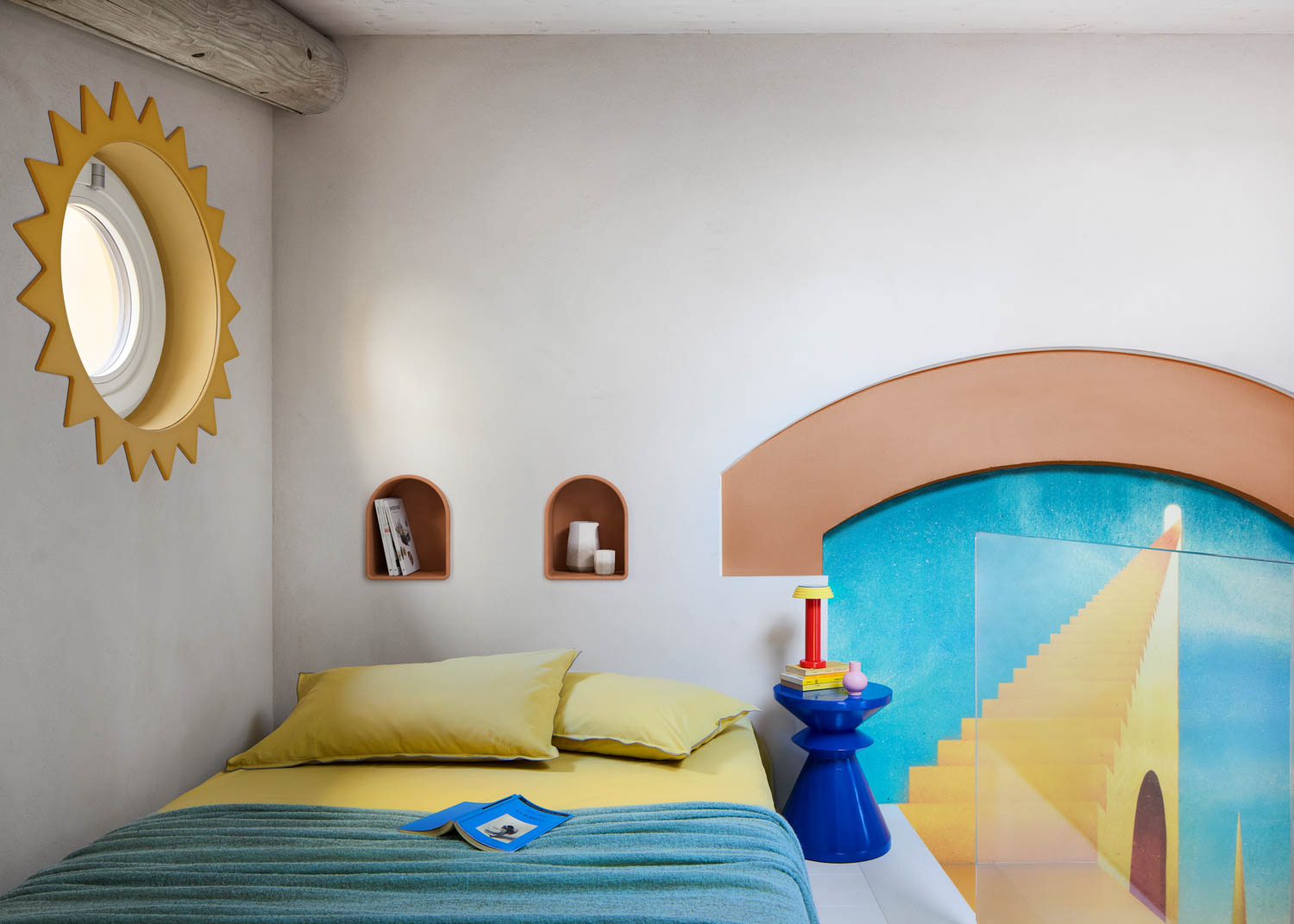 A bedroom with a bed featuring yellow bedding and a blue throw, a sun-shaped window frame, arched wall niches, a blue pedestal side table, and a mural of a staircase.