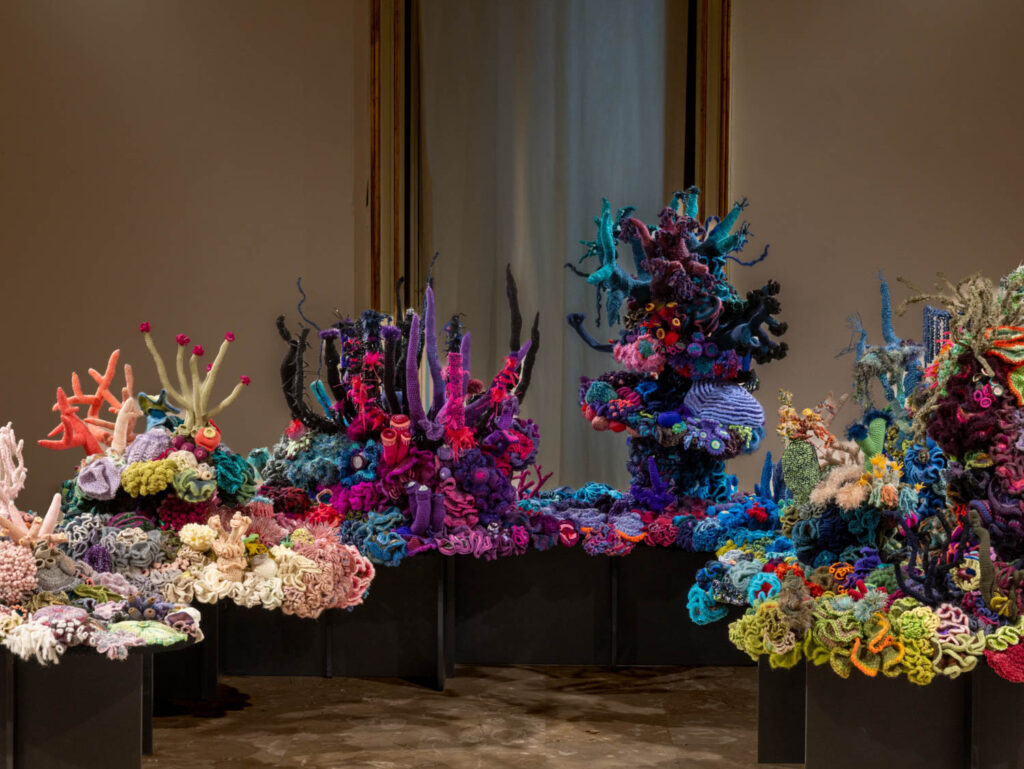 A large, intricate fiber art installation depicting a vibrant coral reef, displayed on several black pedestals.