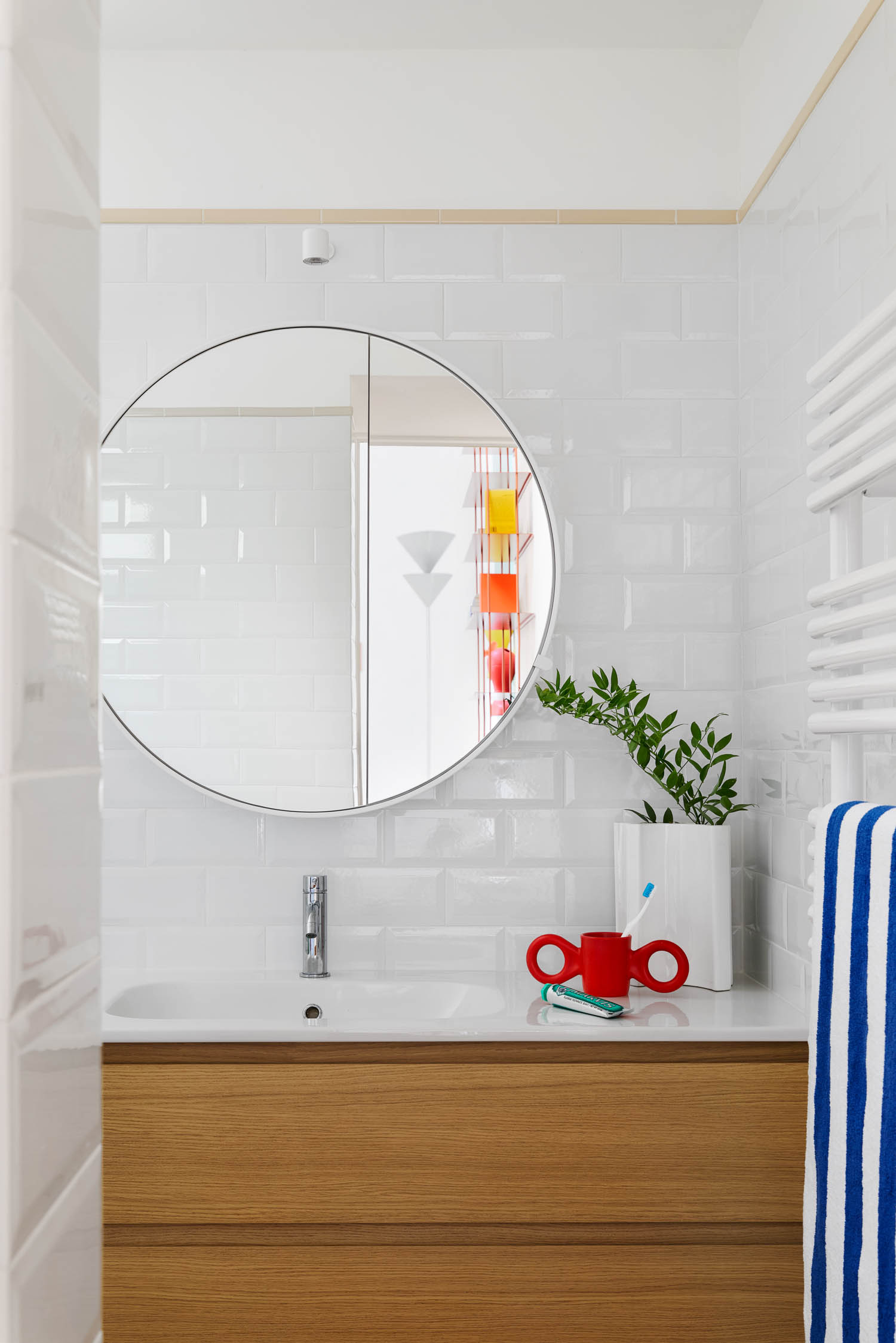Bathroom vanity with a round mirror, white subway tile backsplash, a potted plant, and a red toothbrush holder on the counter.