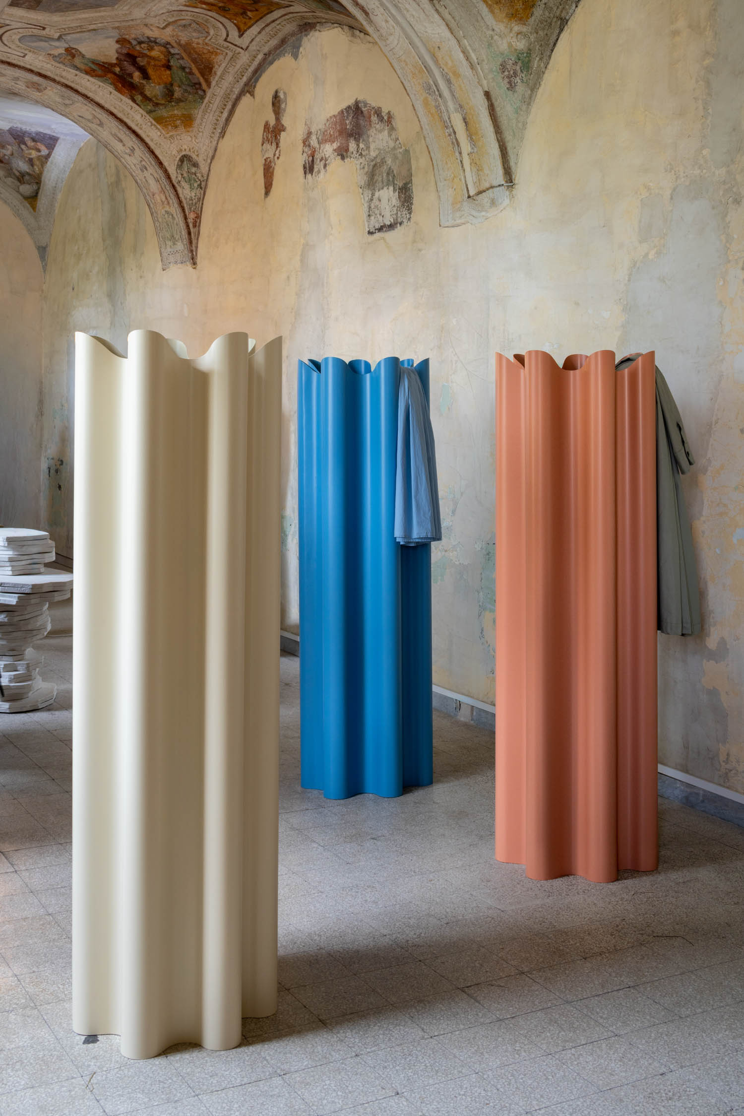 Three fluted, cylindrical coat racks in cream, blue, and terracotta standing in a historic room with frescoed walls.