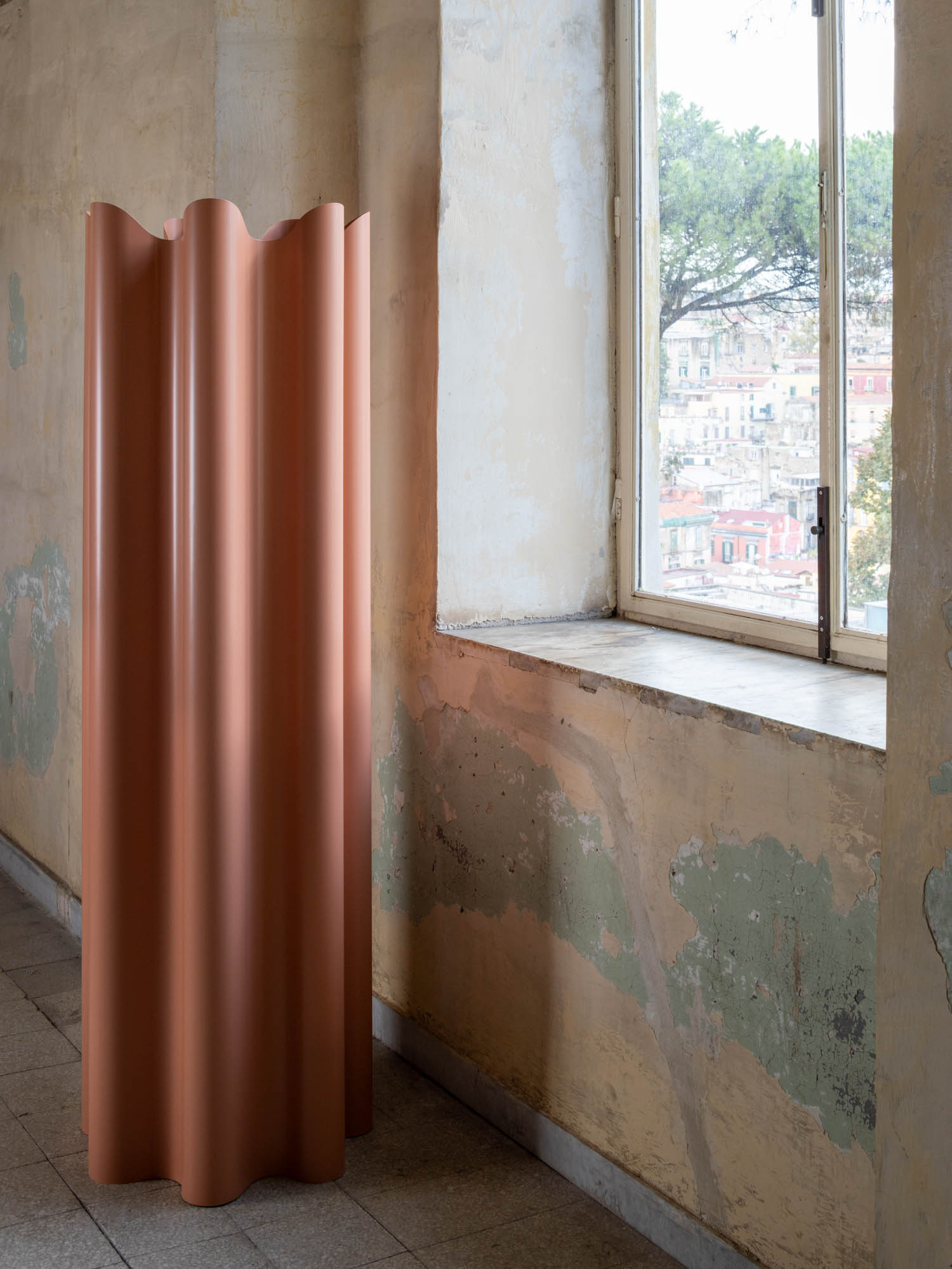 A tall, cylindrical, corrugated pink room divider stands in a room with peeling plaster walls next to a window overlooking a cityscape.