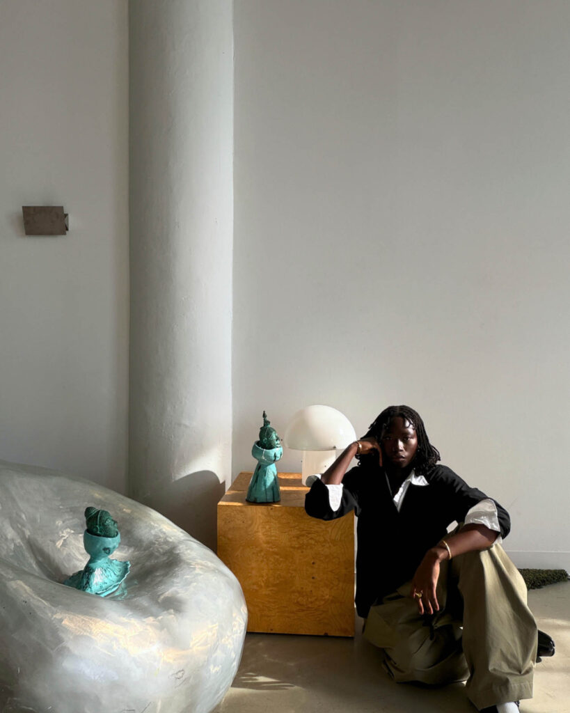 A person with braids sits on the floor next to a yellow wooden pedestal with two small green sculptures and a globe lamp.