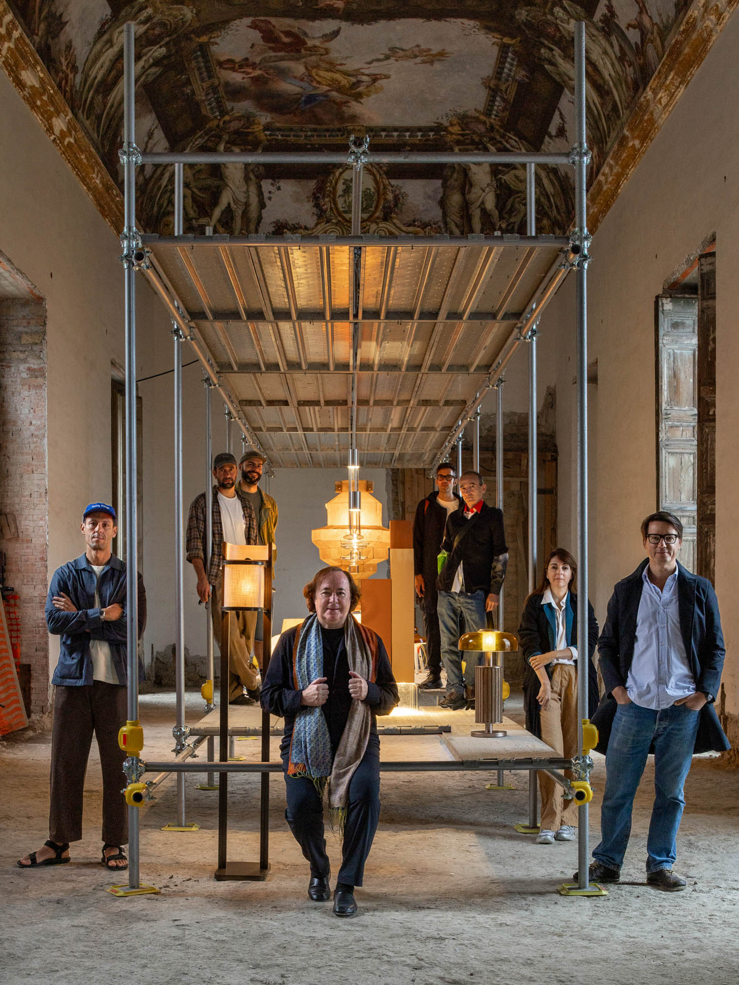 A group of seven people posing around a metal scaffolding structure inside a room with a frescoed ceiling, featuring several lamps displayed on the platform.