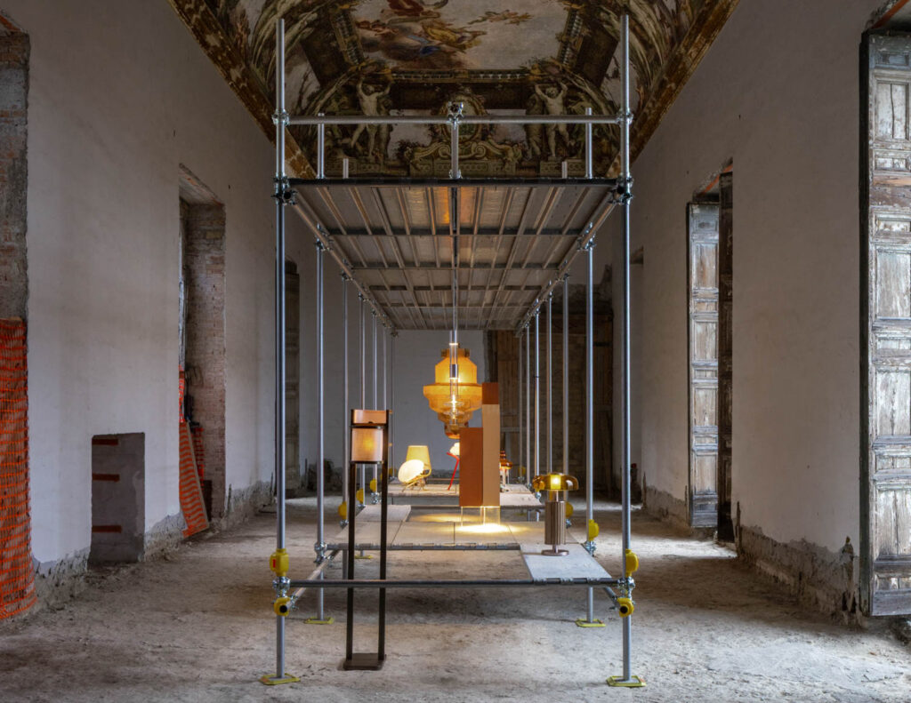 A row of modern lamps displayed on metal scaffolding inside a long, historic room with a vaulted, painted ceiling.