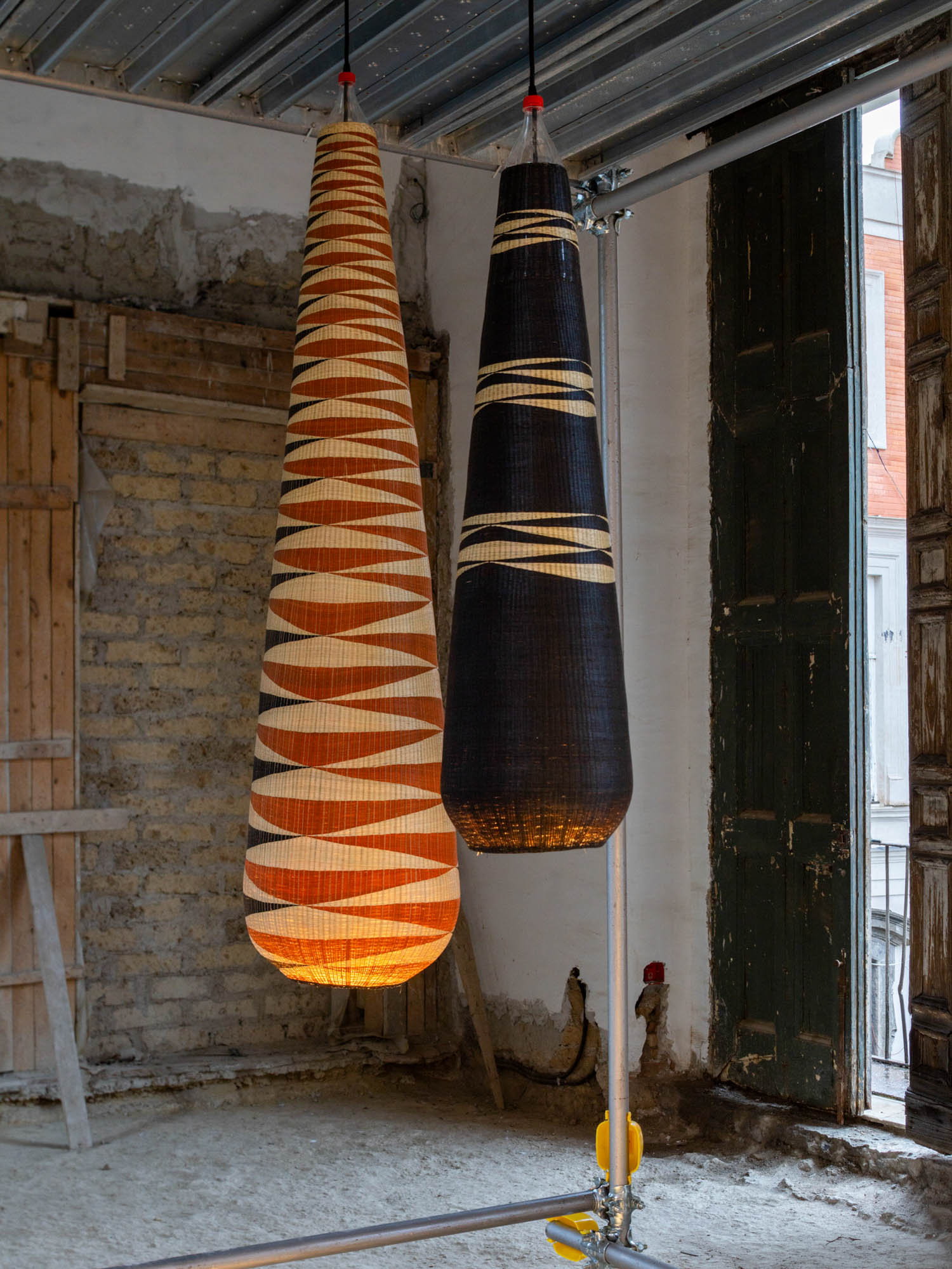 Two tall, teardrop-shaped woven pendant lights, one orange and white and one black and white, suspended in an unfinished room.