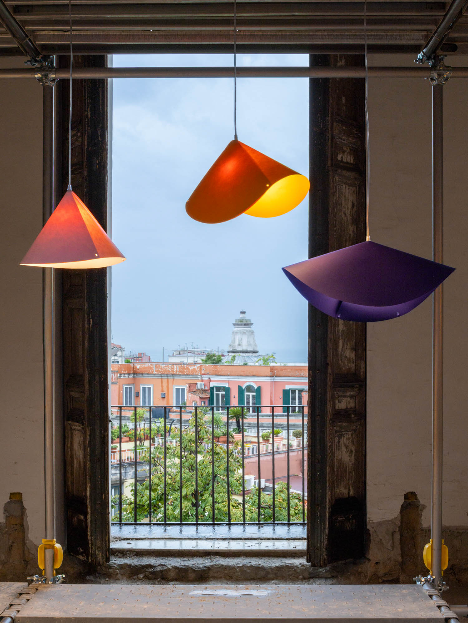 Three cone-shaped pendant lamps in orange, yellow, and purple hang in front of a balcony overlooking a city street.