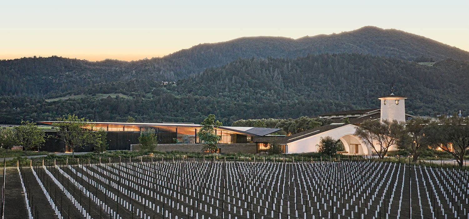 A modern Robert Mondavi winery building with a white tower sits behind rows of young grapevines at the base of a mountain range.