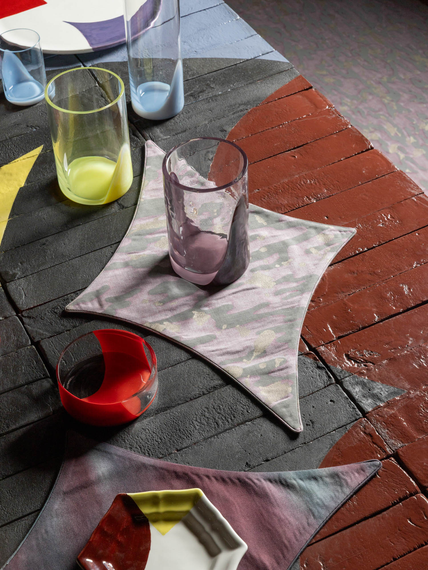Several colorful glass vessels and square fabric coasters arranged on a painted wooden table.