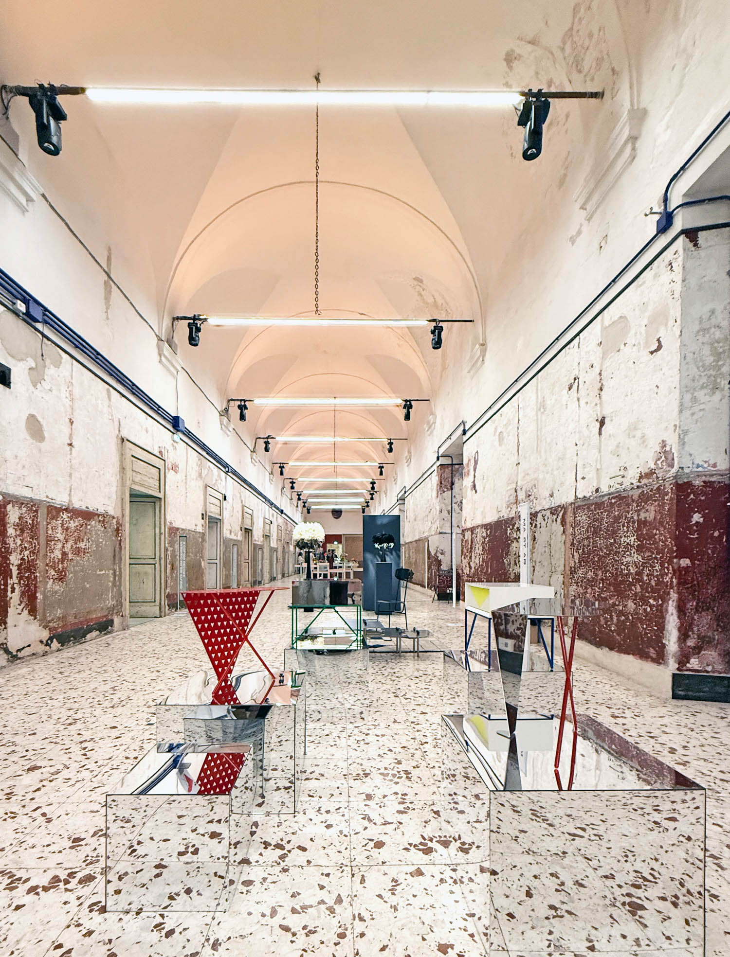 A long, vaulted corridor with weathered walls, displaying abstract furniture and reflective pedestals on a terrazzo floor.