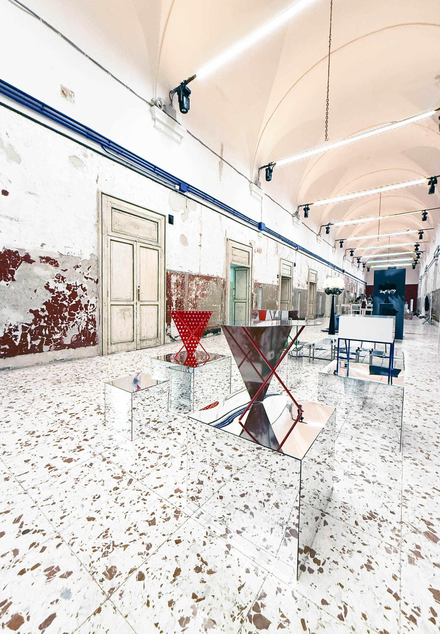 Art installation featuring geometric sculptures displayed on mirrored cubes in a long hall with vaulted ceilings and peeling walls.