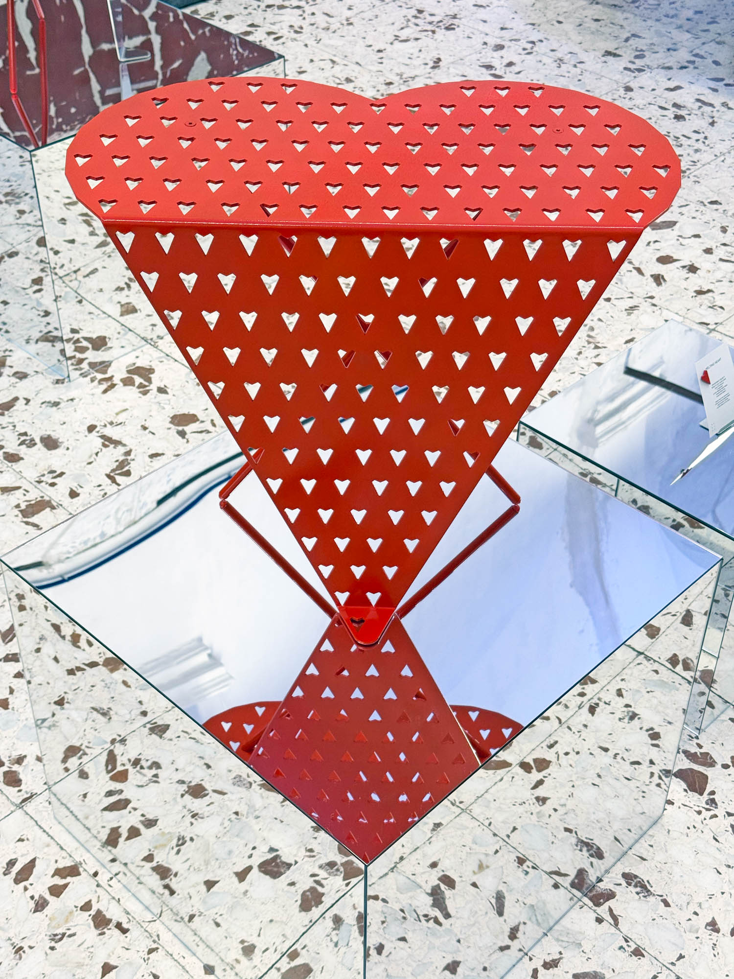 Red heart-shaped metal stool with a perforated heart pattern, resting on a mirrored pedestal.