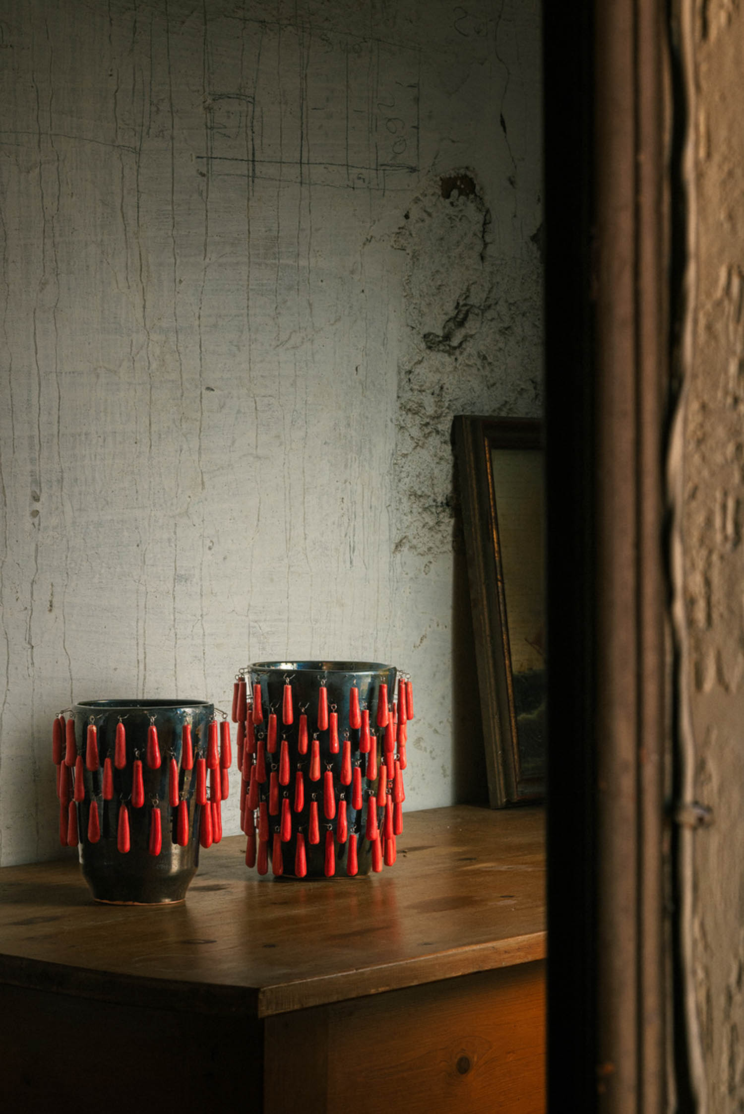 Two dark ceramic vases on a wooden table, both decorated with rows of dangling red, teardrop-shaped charms.