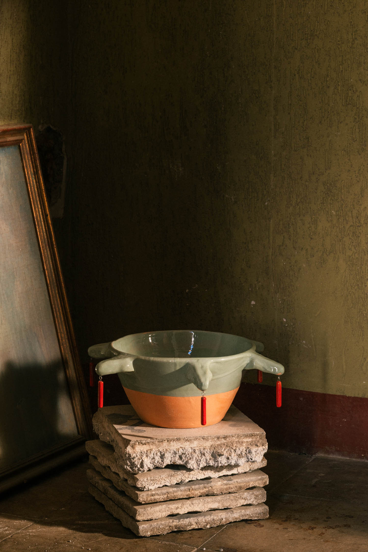 A two-toned ceramic bowl with four protruding handles and small red dangling charms, resting on a stack of stone tiles.