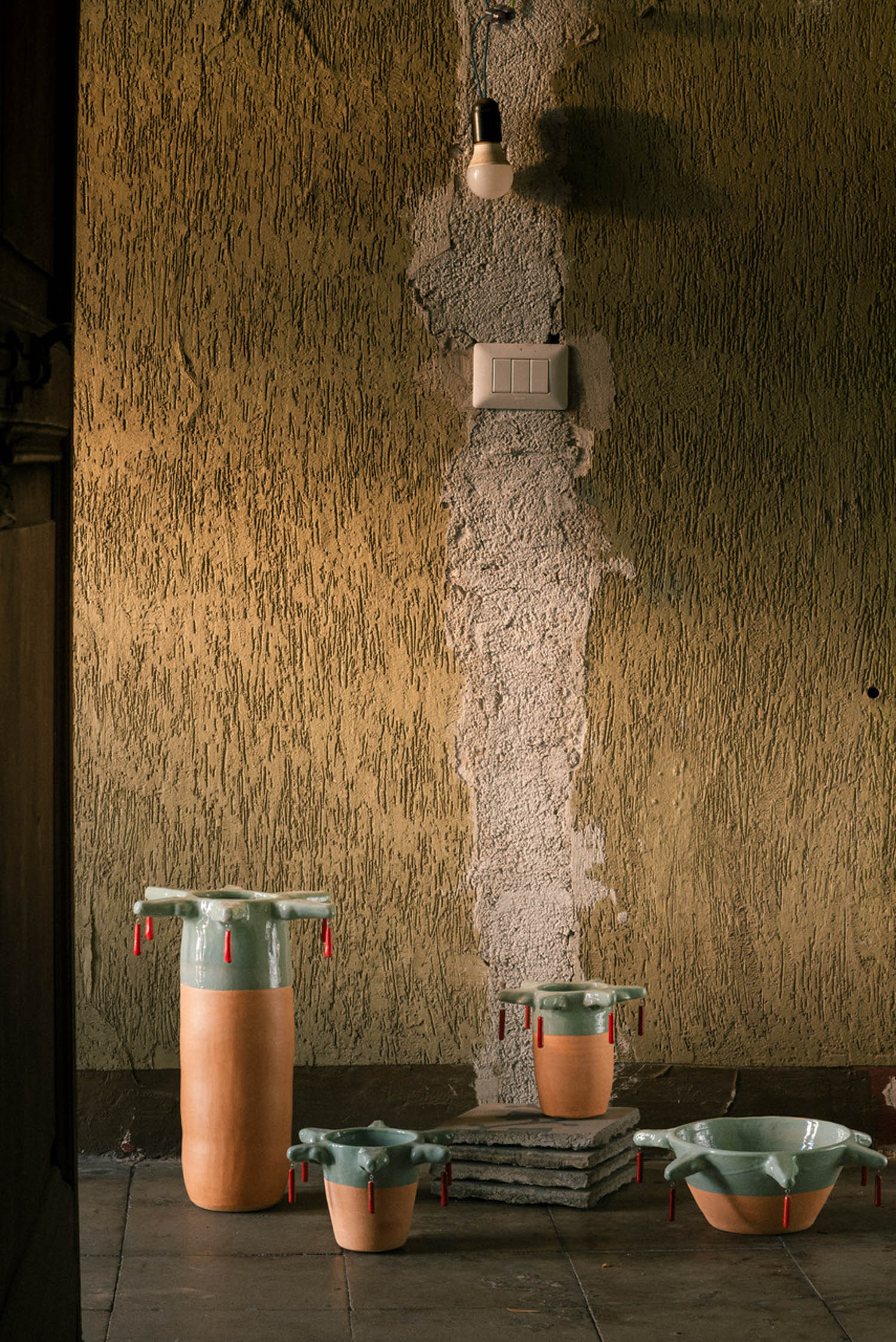 Four ceramic vessels with light green glazed rims and dangling red accents, arranged on a stone floor against a textured wall.