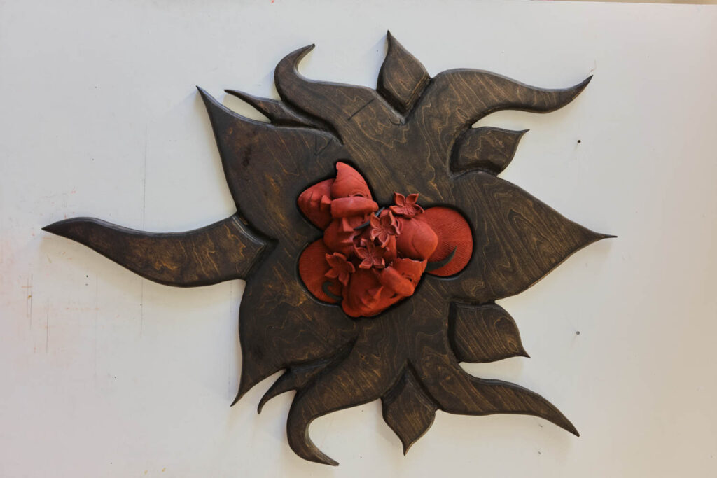 A dark, stained-wood sun-shaped plaque featuring a central red sculpture of an anatomical heart surrounded by small flowers.
