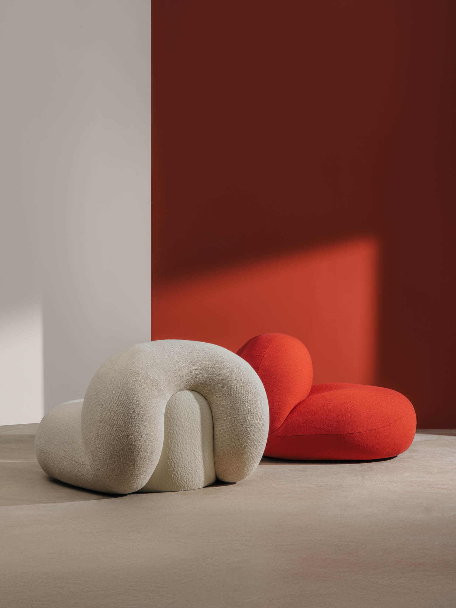 A white and a red modern lounge chair sit on a light floor against a two-tone wall, with natural light casting shadows.