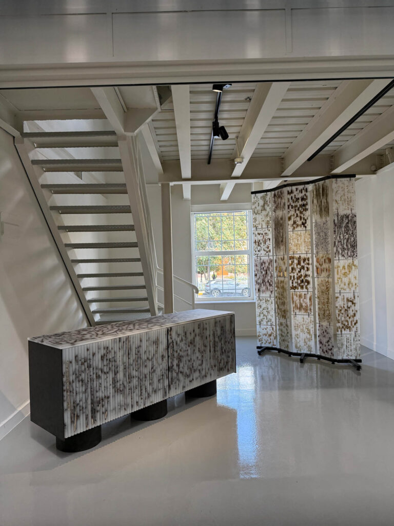 A modern console table and a folding room divider, both featuring vertical patterns of pressed organic material, sit in a bright room with an open staircase.