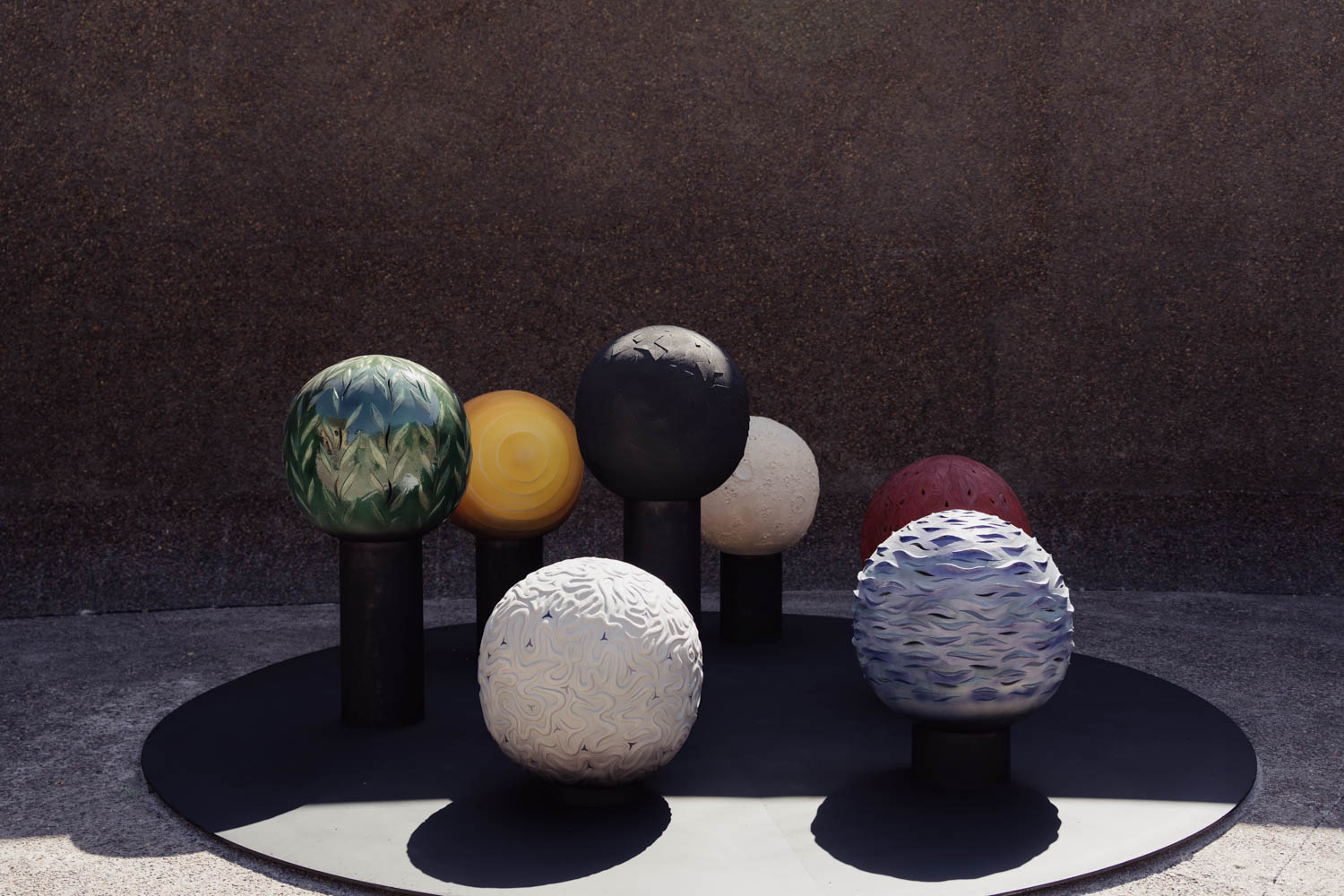 Seven sculptural spheres with varied patterns and colors, each mounted on a short pedestal, arranged on a circular platform.
