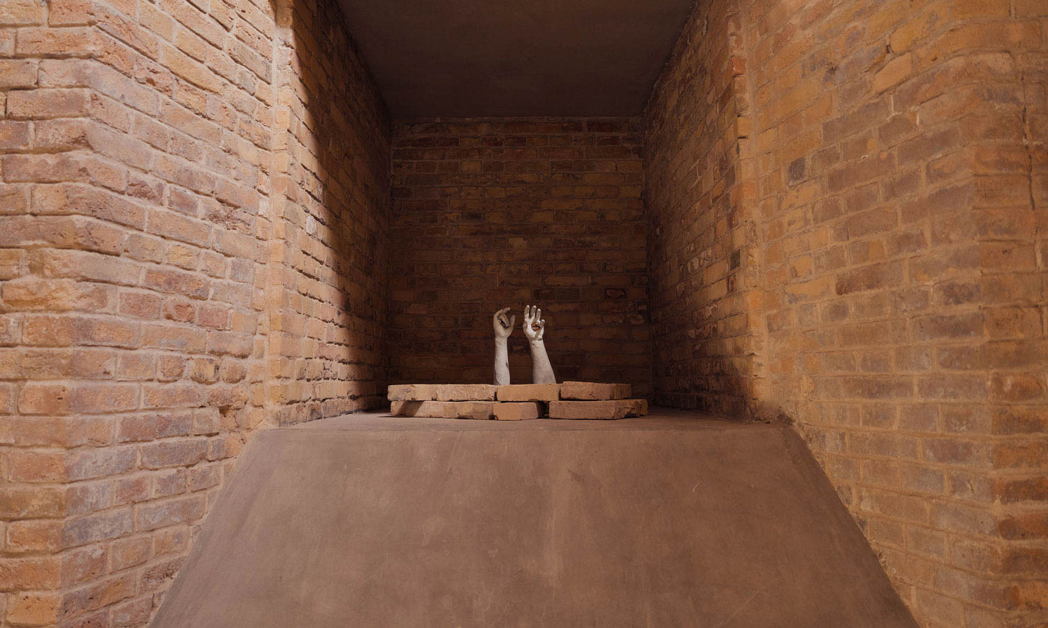 Two sculptural hands emerge from behind a row of bricks within a brick-walled alcove.
