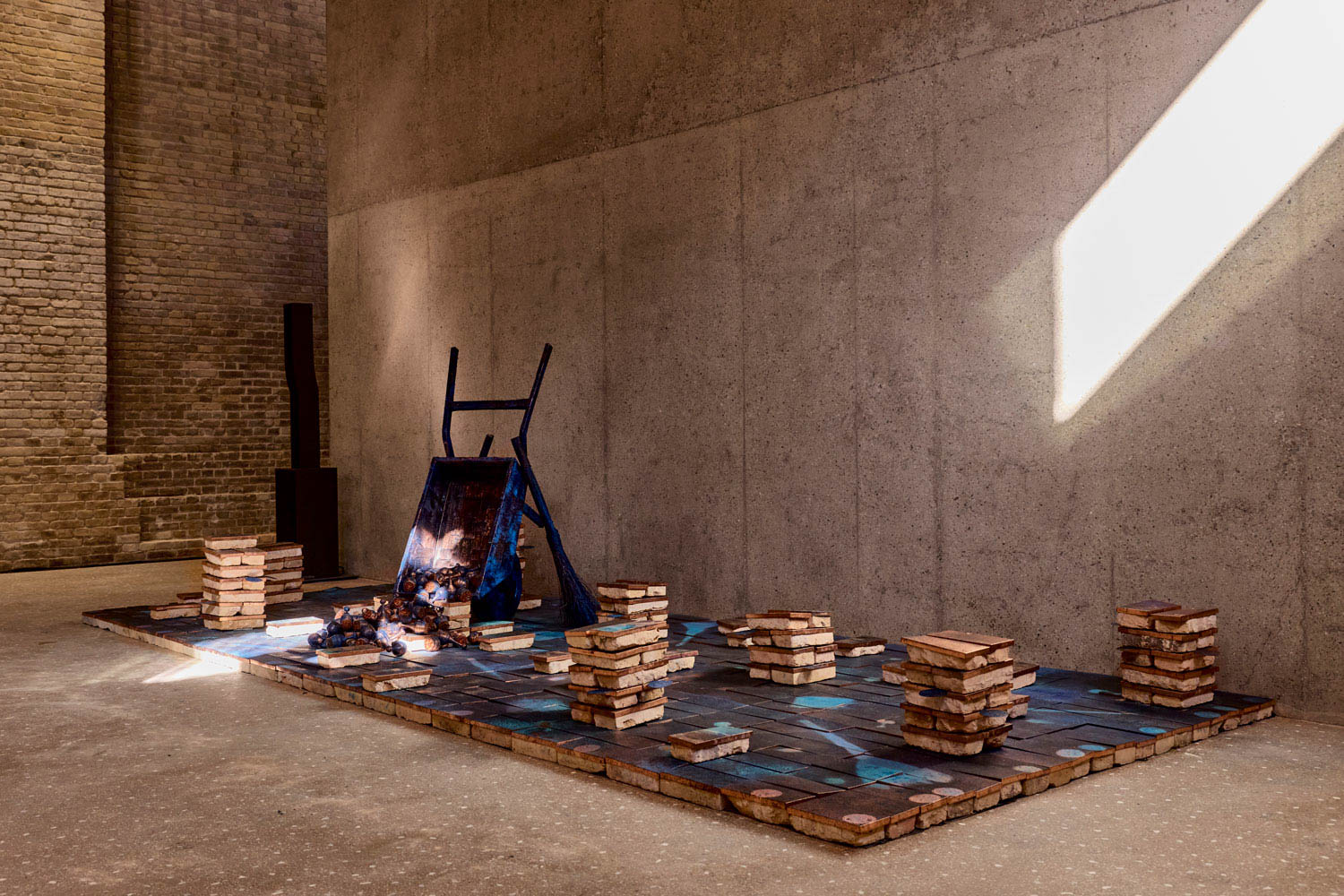An art installation featuring an overturned blue wheelbarrow surrounded by stacks of bricks on a tiled floor.