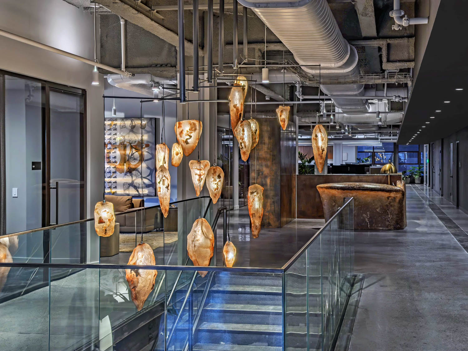 A group of organic-shaped, glowing pendant lights suspended in an office hallway above a glass-railed staircase.