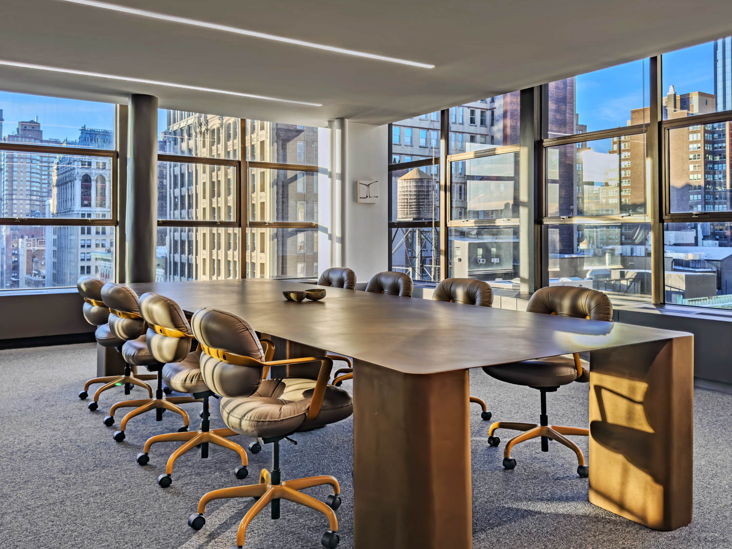 A long, metallic boardroom table surrounded by eight rolling chairs in a room with floor-to-ceiling windows overlooking a city skyline.