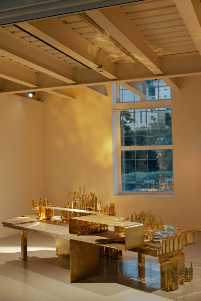 A brass sculpture resembling an abstract cityscape sits on a wooden table in a gallery space near a window.