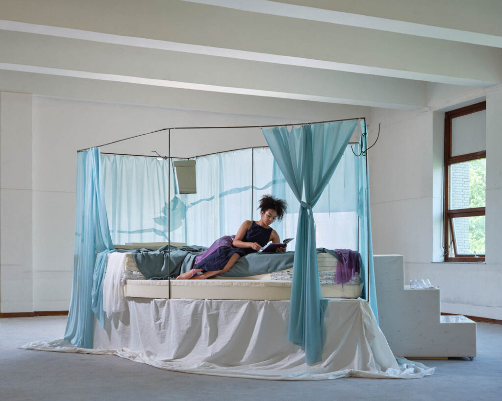 A person sits on a bed frame draped in light blue fabric, reading a book.