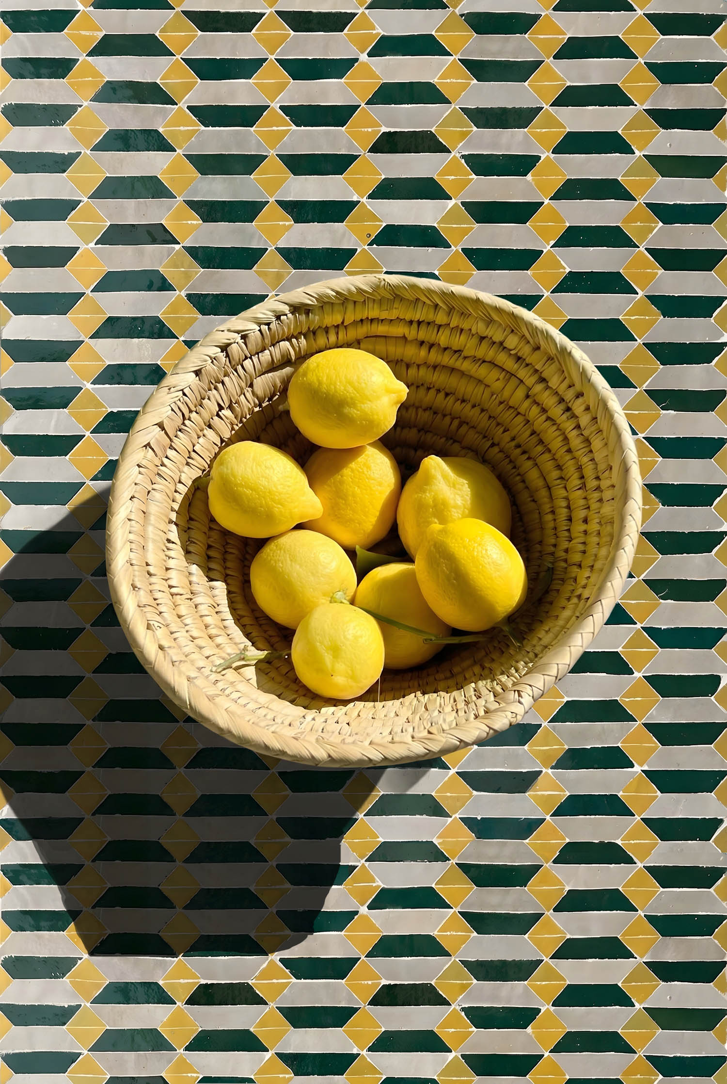 A woven basket with yellow lemons sits on a patterned geometric tile surface, casting a shadow to the left.