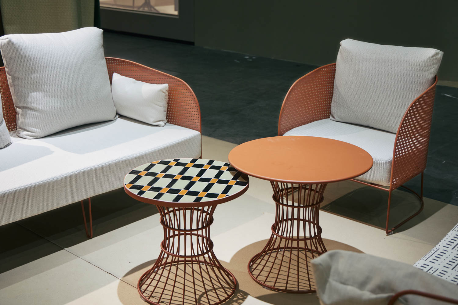 Two metal-framed chairs with white cushions and two round tables, one with a checkered top, are arranged on a light-colored floor against a dark background.