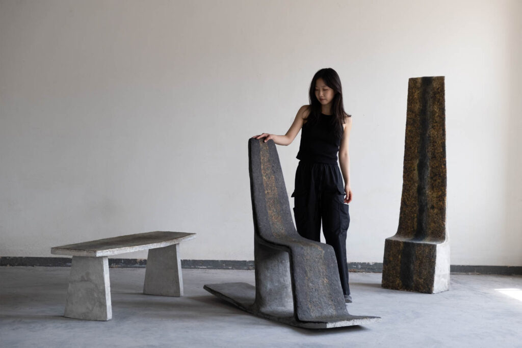 A person stands beside a sculptural chair, with a bench and a tall, narrow pillar-like piece nearby, all featuring a textured, weathered finish.