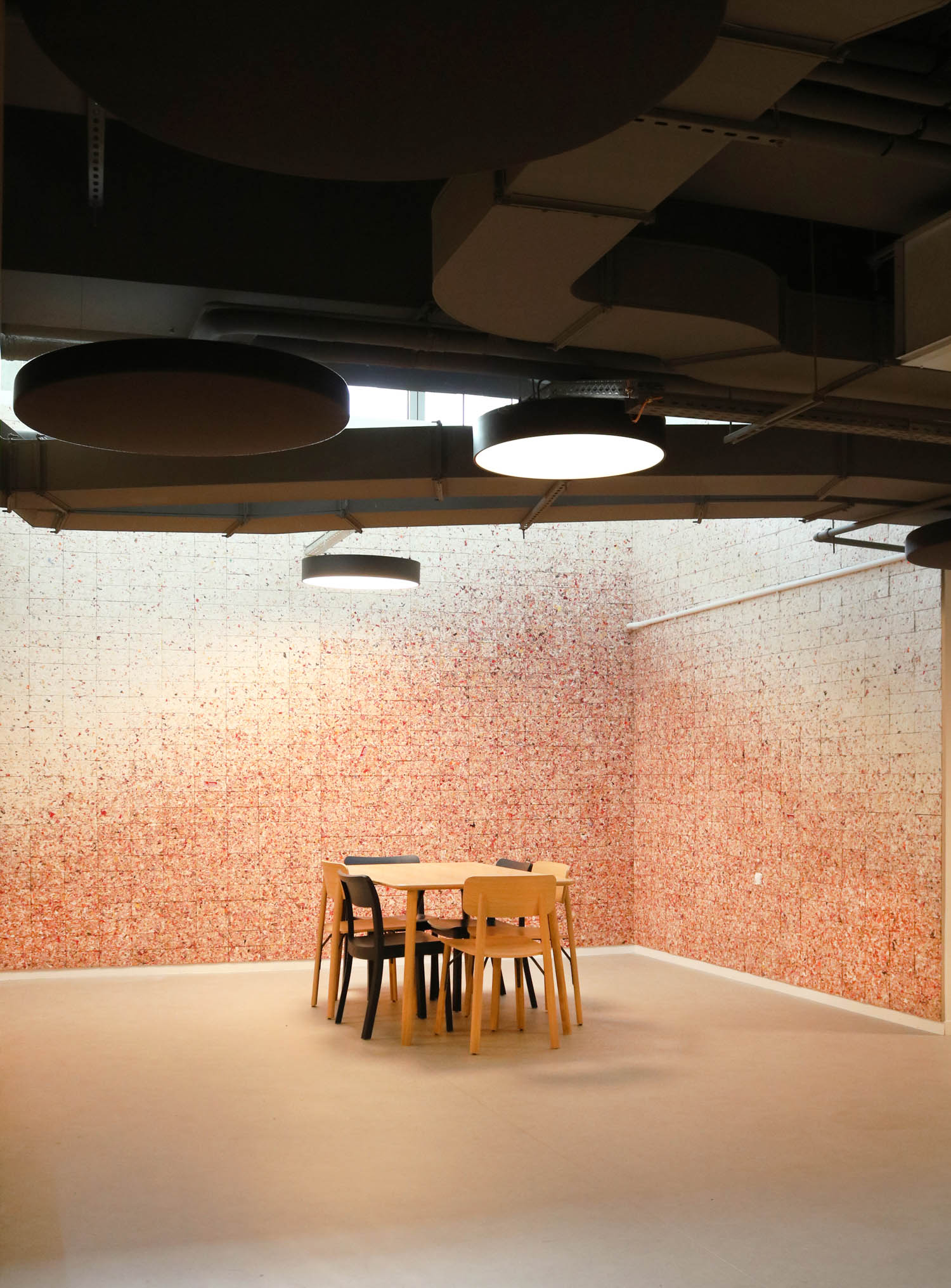 A round table with four chairs centered in a room with textured, pink-speckled walls and suspended light fixtures.