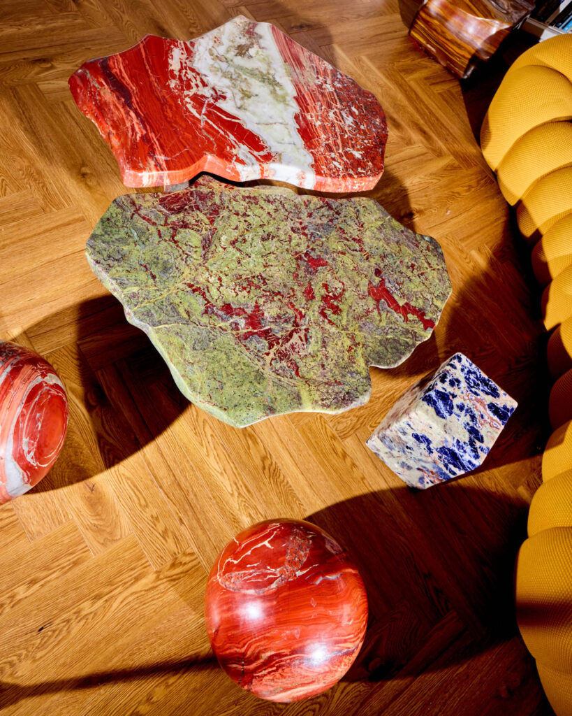 Two organic-shaped marble table tops and two stone spheres in red and green, alongside a blue and white cube, arranged on a wood floor next to a yellow sofa.