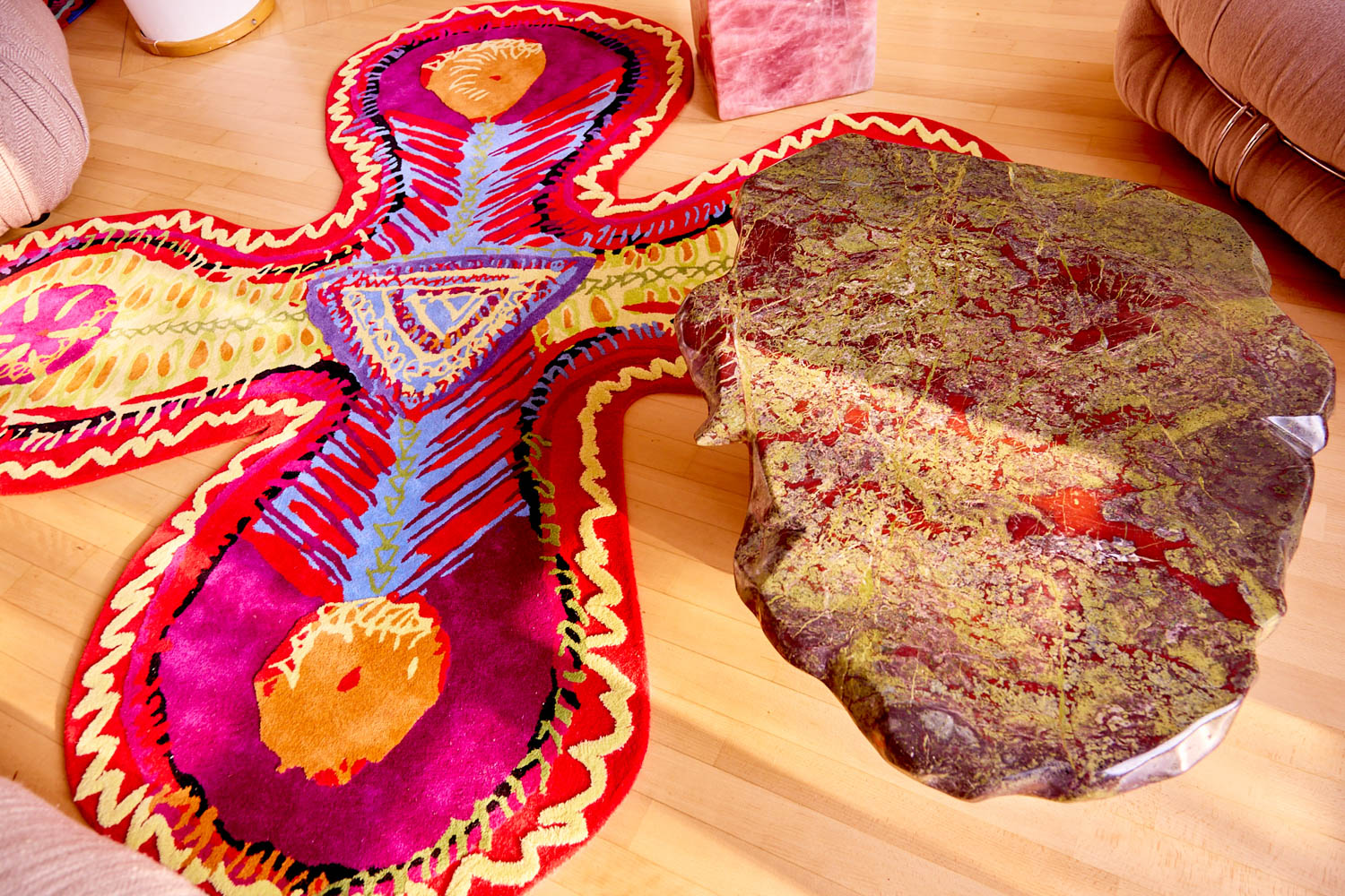 An abstract, irregular-shaped area rug with red, purple, and yellow patterns beside a table with a mottled, rock-like surface.