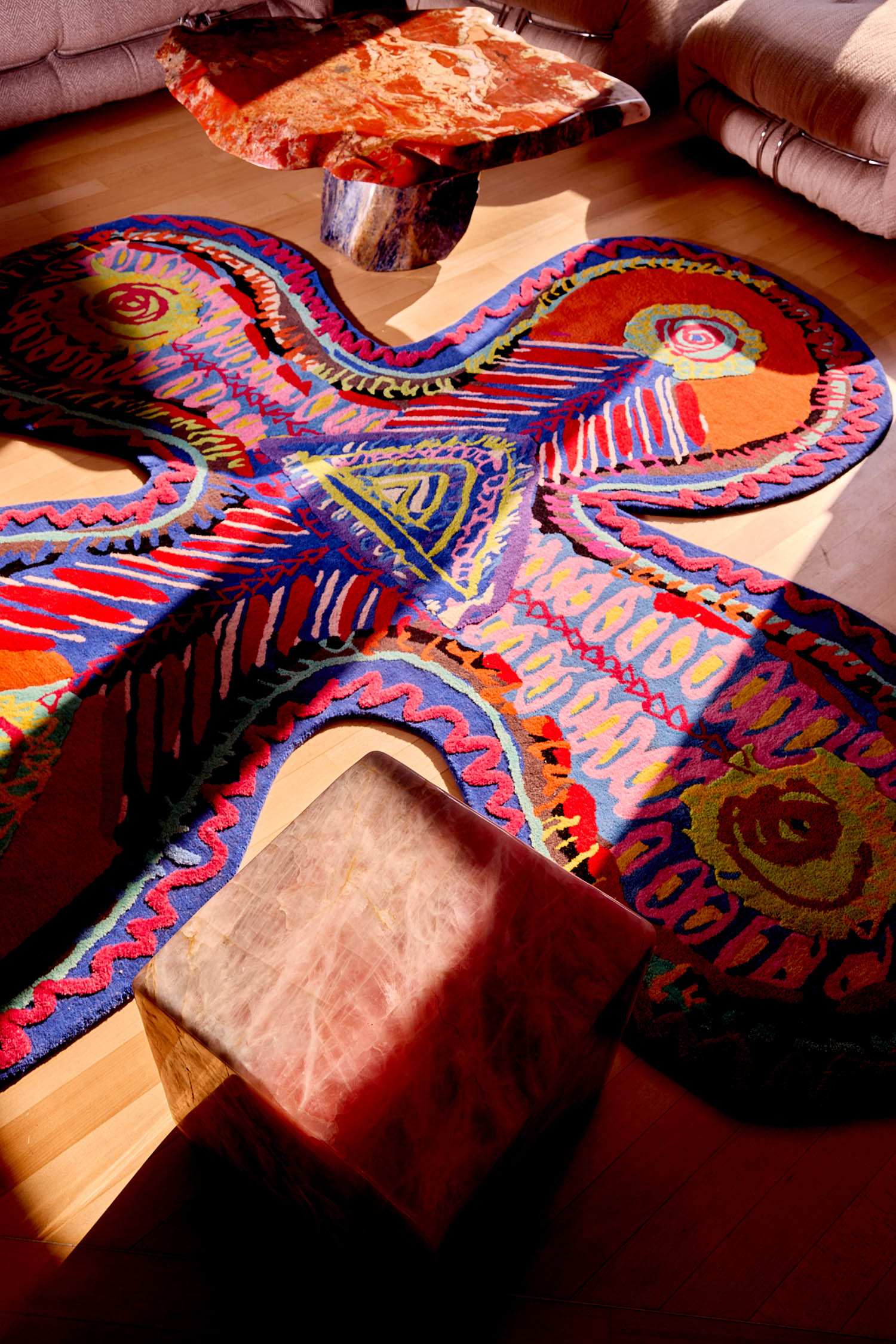 An irregularly shaped, colorful tufted rug with abstract patterns on a wood floor, surrounded by a stone coffee table and a square stone stool.