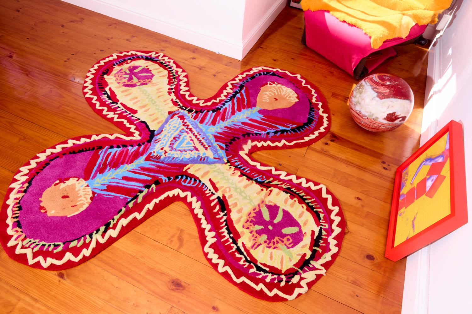 An abstract, cross-shaped rug with magenta, yellow, and blue patterns on a hardwood floor, next to a globe and a framed piece of art.