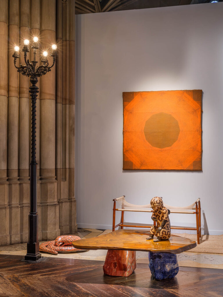 A gallery interior featuring an orange abstract painting on the wall, a wooden coffee table with a small patterned figure, a low bench, and an ornate floor lamp.