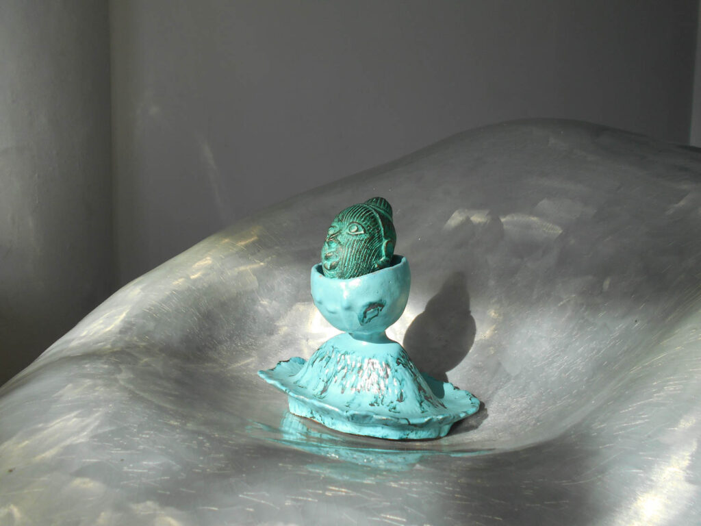 A small, green sculpted head resting inside a turquoise, pedestal-shaped ceramic bowl set on a metallic, curved surface.
