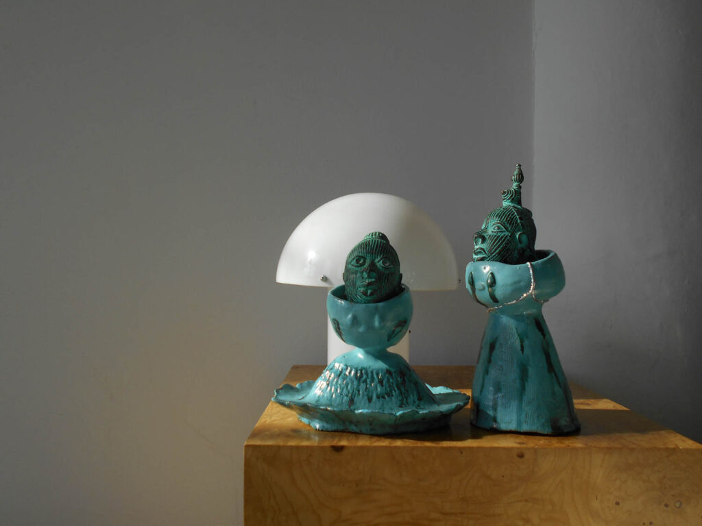 Two teal-colored ceramic sculptures of heads resting in bowl-like forms, positioned on a wooden surface in front of a white dome lamp.