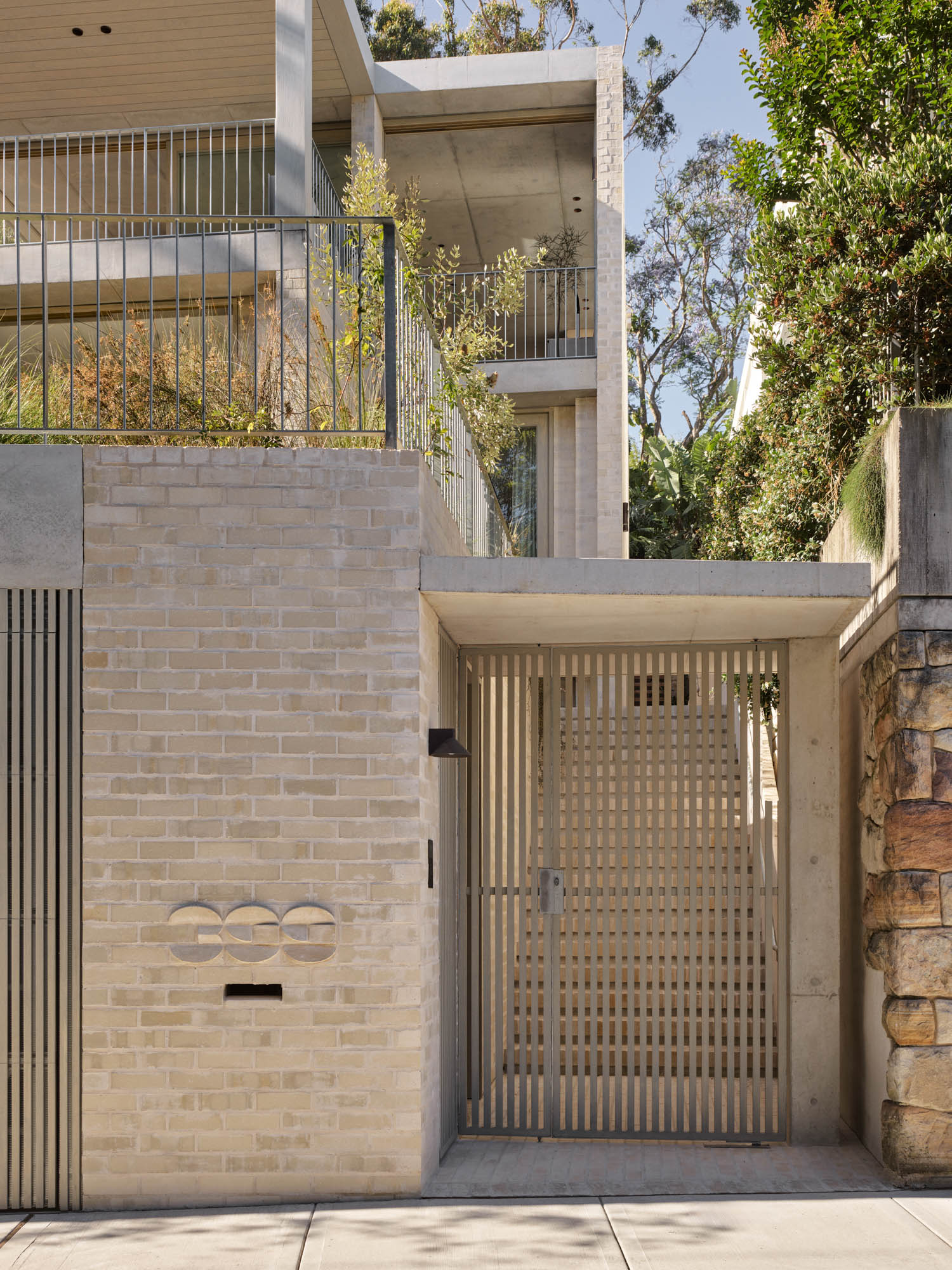Modern beige brick residence with a metal gate and horizontal slats, surrounded by greenery and stonework, seen from the sidewalk.