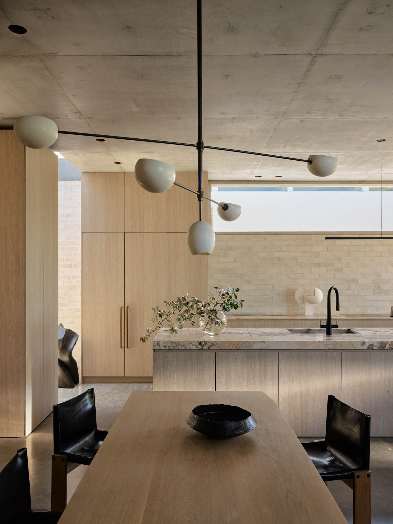 Modern kitchen and dining area with light wood cabinetry, marble island, black faucet, minimalist light fixture, black chairs, and a decorative bowl on the table.