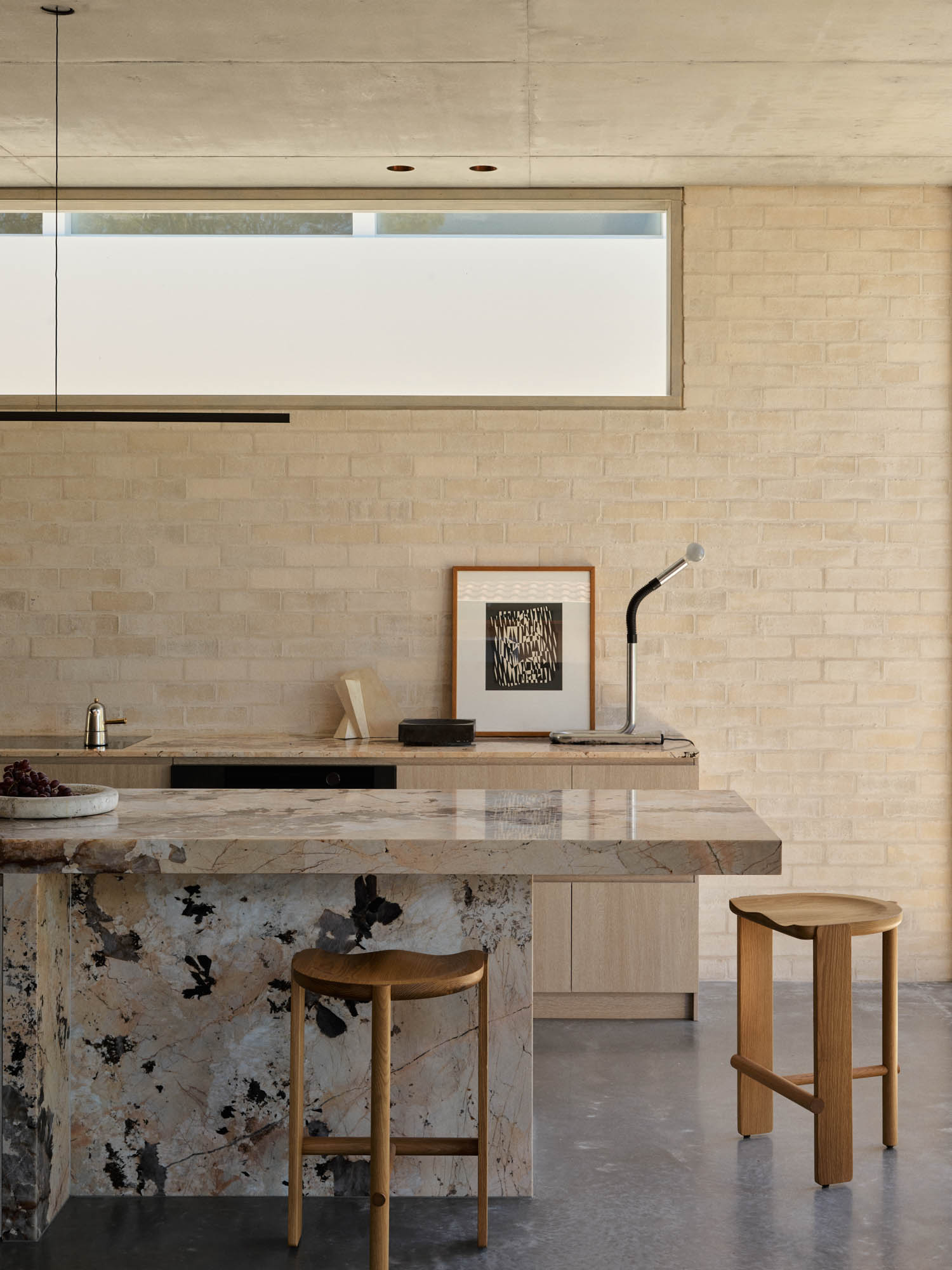 Modern kitchen with a marble island, two wooden stools, built-in sink, wall art, and a lamp on a light brick wall under a horizontal window.