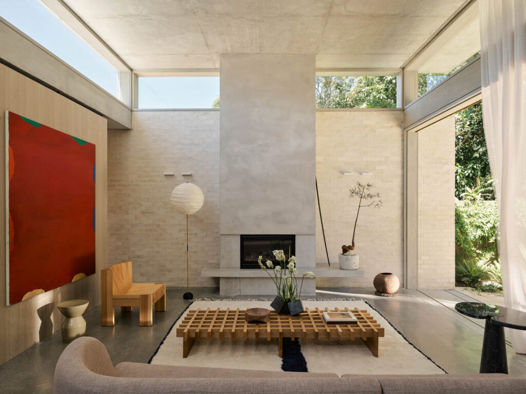 Modern living room with a concrete fireplace, minimalist furniture, large windows, abstract red art, and a coffee table with books and flowers. Natural light fills the space.