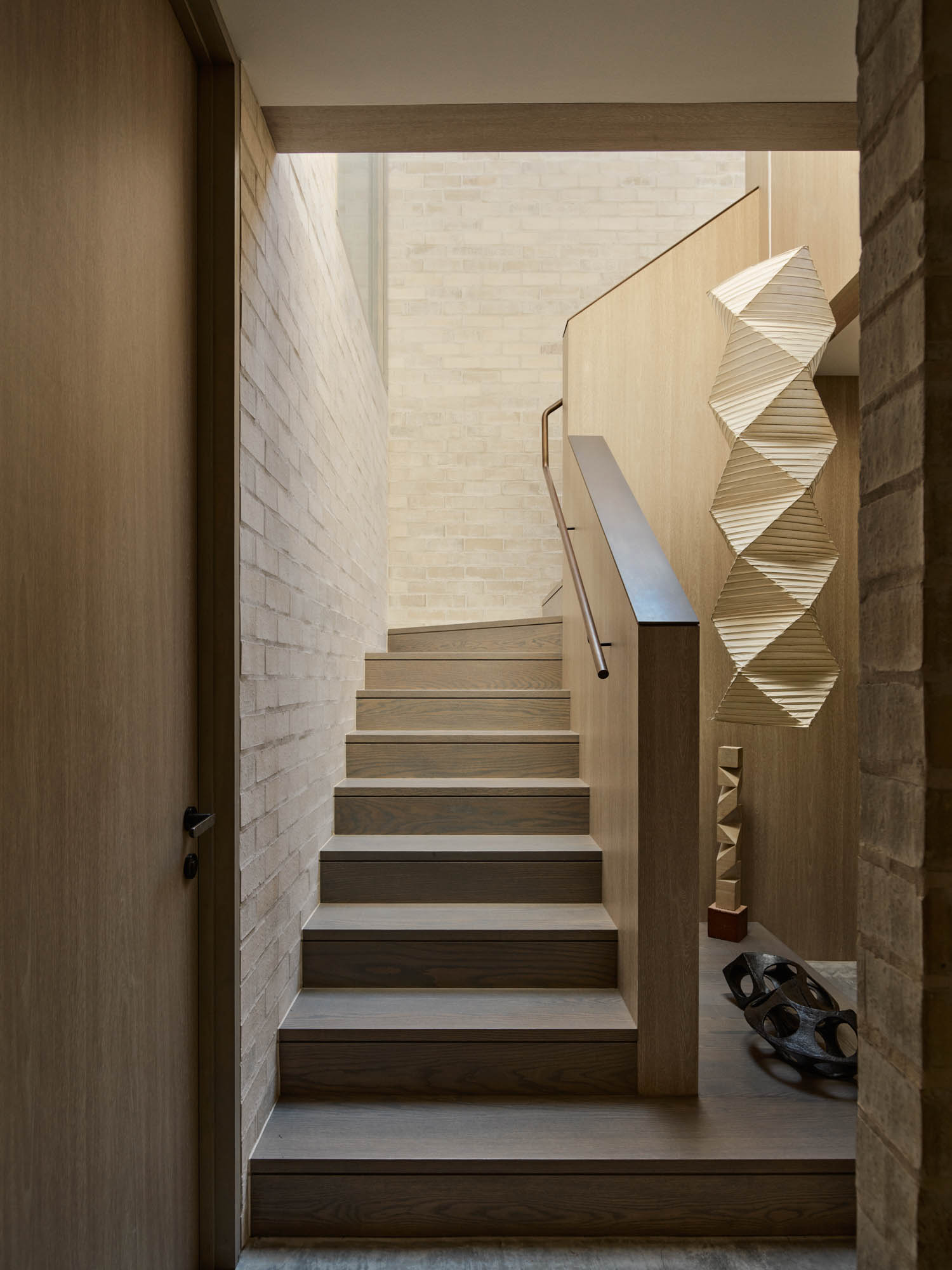 Wooden staircase with a simple handrail, beige brick walls, geometric hanging sculpture, and decorative objects on the floor in a modern interior.