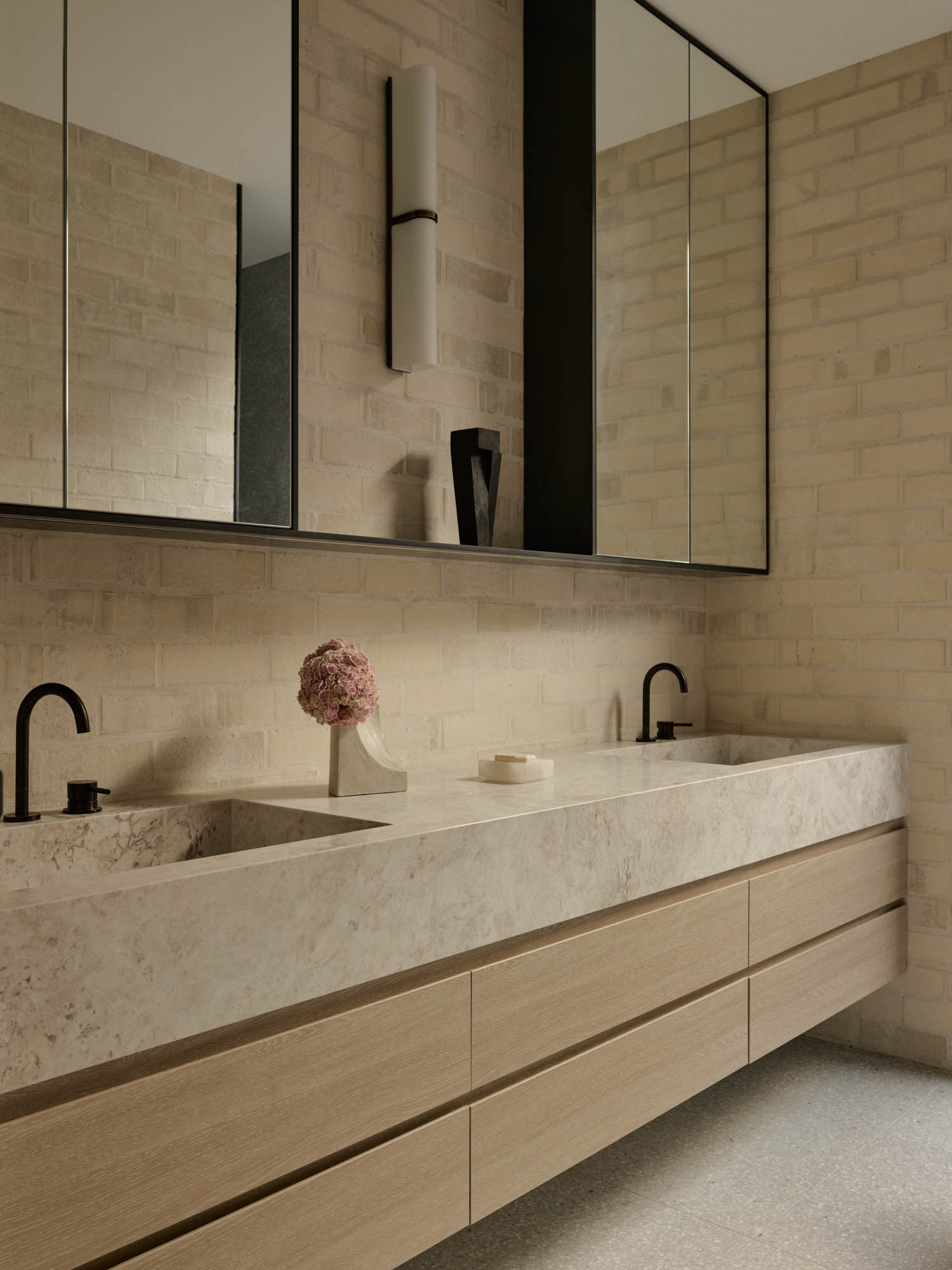 Modern bathroom with beige stone countertop, dual sinks, black faucets, light wood cabinetry, large mirrors, and a single pink flower in a vase. Cream brick walls and minimalist decor.