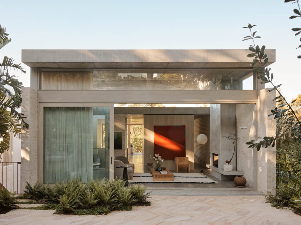 Modern single-story house with large glass windows, minimalist decor, and plants in the front yard. Interior features neutral tones, a red accent wall, and contemporary furnishings.