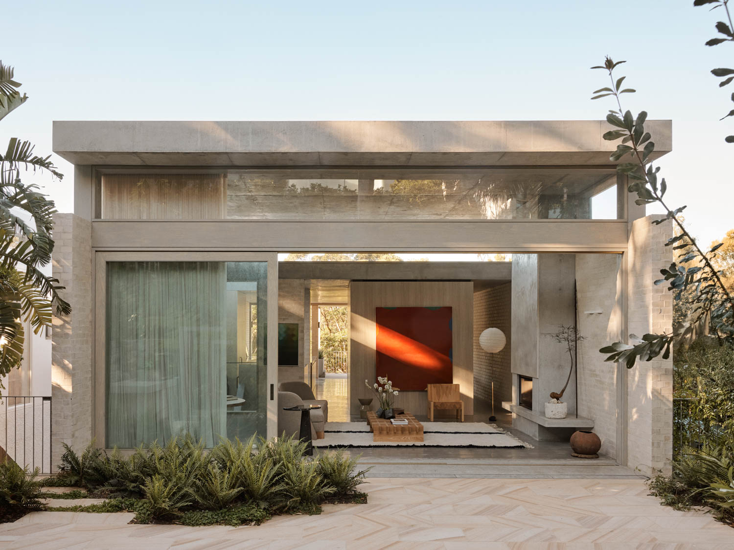 Modern single-story house with large glass windows, minimalist decor, and plants in the front yard. Interior features neutral tones, a red accent wall, and contemporary furnishings.